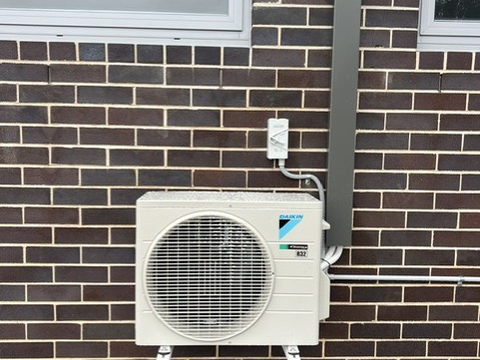 Outdoor air conditioning unit on brackets against a brick wall, with pipework running beside a window.